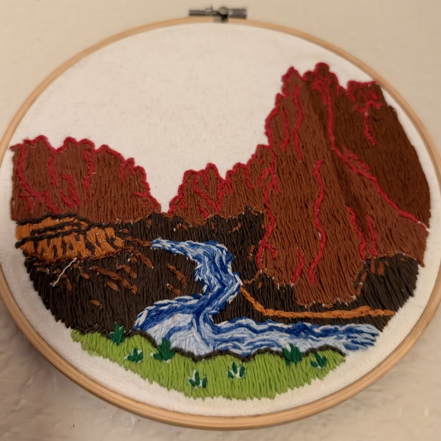 Hand embroidery of red canyon cliffs above a blue river, in a wooden hoop