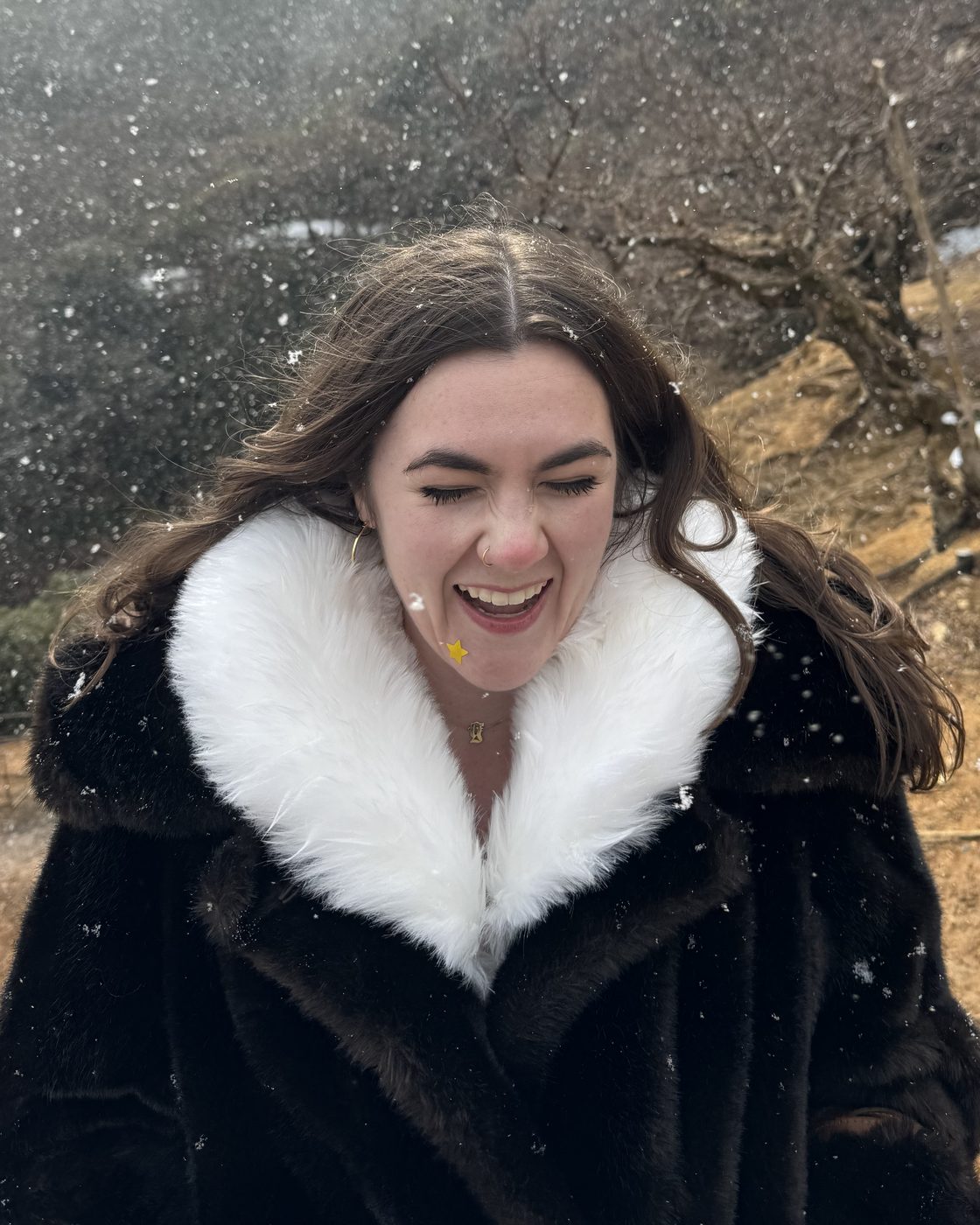 Ivey laughing in a black coat with a white fur collar as snow falls