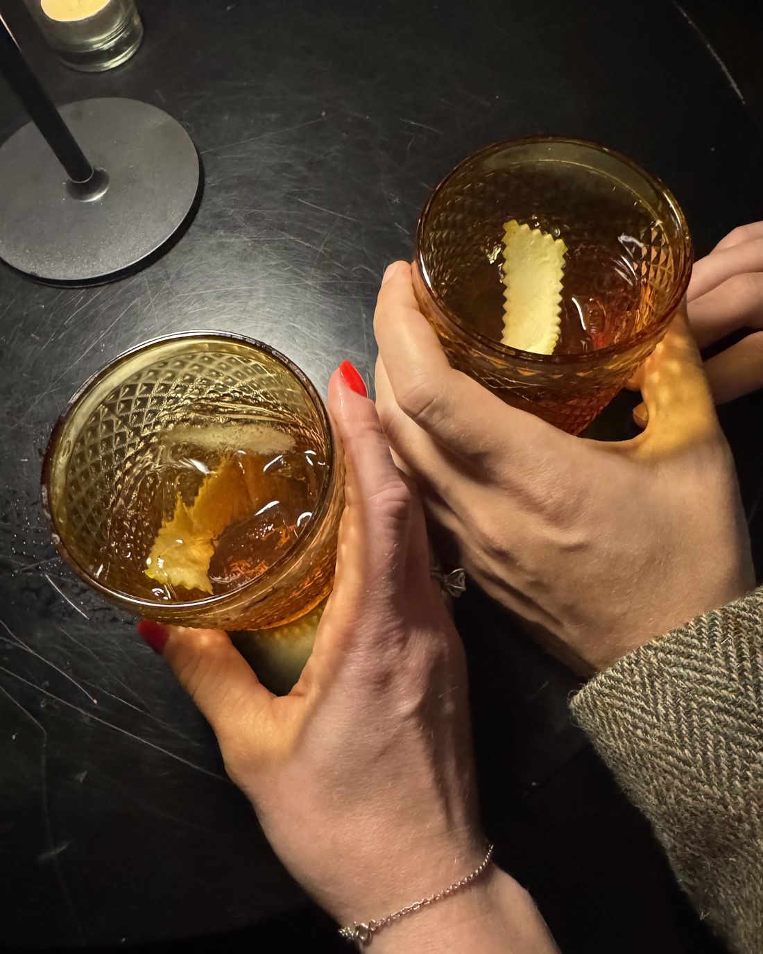 Two hands clinking amber cocktails on a dark table
